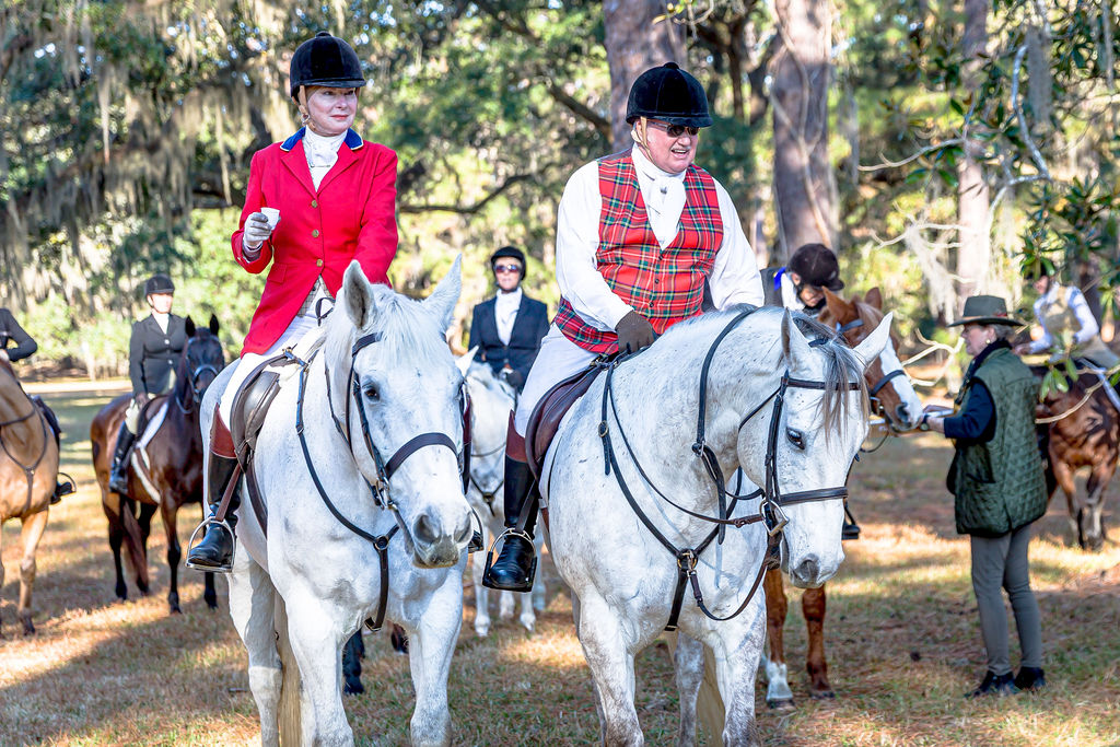 Two mounted riders in traditional hunt attire gathered with the field beneath live oaks.