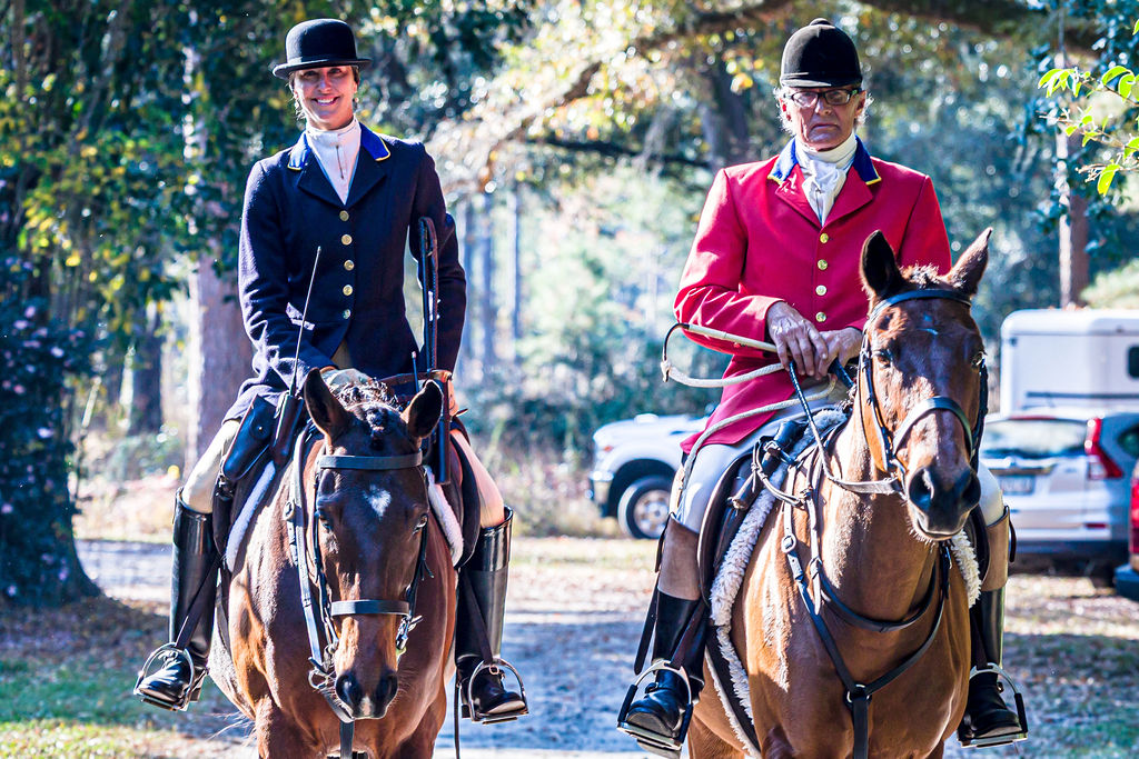 Two mounted riders in formal hunt coats at a woodland meet.