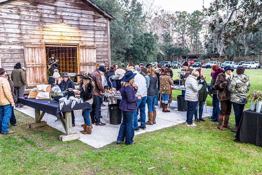Members and guests gathered around an outdoor hunt breakfast beside a weathered wooden building.