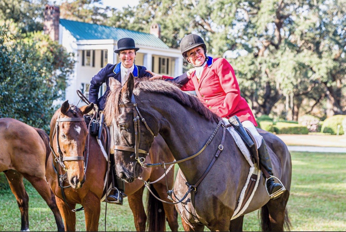 Two mounted members, one in a scarlet coat, drawn up together on the lawn before a white plantation house.
