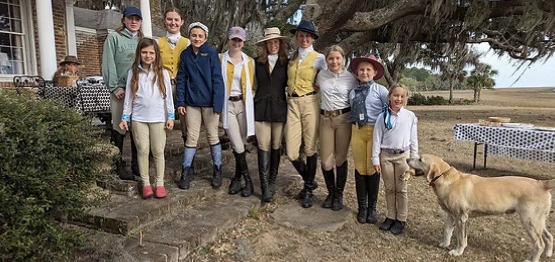 A wide group of junior riders in front of a house, with a dog among them.
