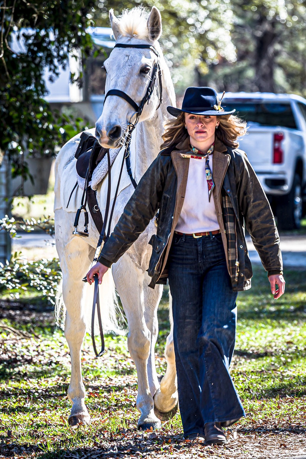 An equestrian in a broad-brimmed hat leading a saddled white horse through dappled sunlight.