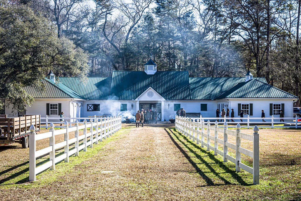 A white stable with a green roof seen along a fenced lane in winter sunlight.