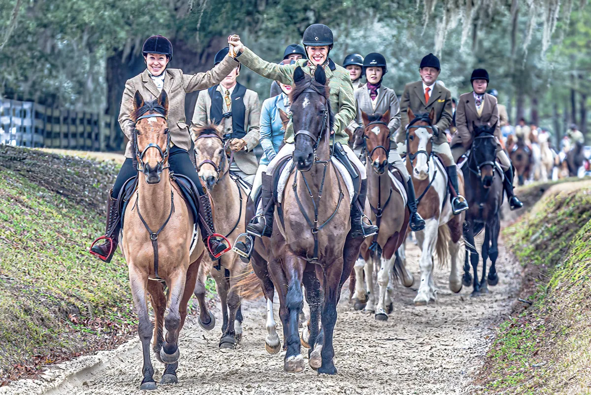 Two tweed-clad riders raising clasped hands in a sunken lane, with a mounted line behind them.